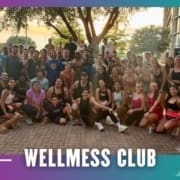 A large group of people pose together outdoors, many smiling and wearing athletic clothes, with trees and buildings in the background. Text at the bottom reads "WELLMESS CLUB" on a colorful border—part of a Buda Running Club group run. Austin Marathon Half Marathon & 5K