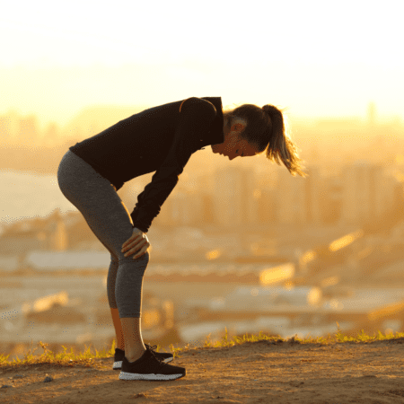 A woman in athletic wear bends over to catch her breath after exercising outdoors on a hill at sunset, finding running motivation as she gazes at the cityscape and water in the background. Austin Marathon Half Marathon & 5K