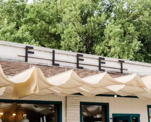 A building with large black letters spelling "FLEET" on the roof, light fabric awnings, teal window frames, and green trees in the background—one of the best coffee shops in Austin for post-run meetups. Austin Marathon Half Marathon & 5K
