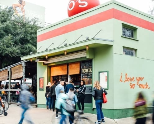 People walk and cycle past a green building with a red “O’s” sign and “i love you so much.” painted on the side—a favorite for post-run meetups and one of the best coffee shops in Austin, with outdoor seating and a welcoming vibe. Austin Marathon Half Marathon & 5K