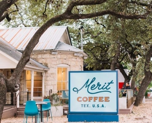 A cozy coffee shop with a stone exterior and a large "Merit Coffee TEX. U.S.A." sign stands under leafy trees—one of the best coffee shops in Austin, perfect for post-run meetups at the teal chairs on the leaf-strewn ground. Austin Marathon Half Marathon & 5K